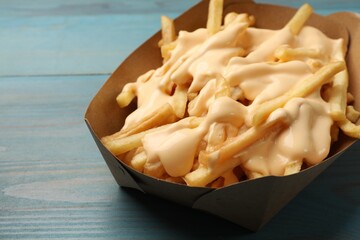 Tasty potato fries and cheese sauce in paper container on light blue wooden table, closeup