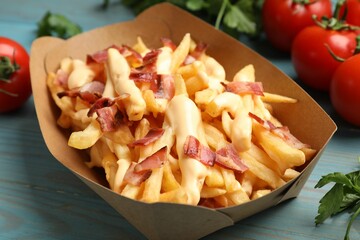 Tasty potato fries, cheese sauce, bacon and products in paper container on light blue wooden table, closeup