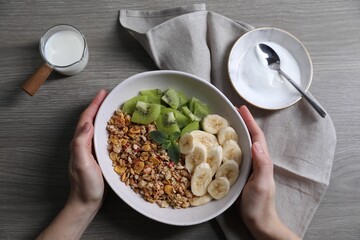 Woman holding bowl of tasty granola with banana and kiwi at grey wooden table, top view