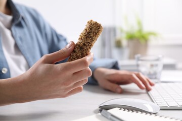 Woman holding tasty granola bar working with computer at light table in office, closeup