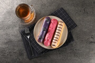 Different tasty glazed eclairs served with tea on grey textured table, flat lay