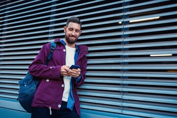 Outdoor fall winter portrait handsome hipster man with beard white t shirt blue shirt maroon jacket holding smartphone ribbed urban wall background - Powered by Adobe