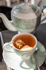 Cup of herbal tea on a matching saucer with teabag and slice of lemon next to green leaf print teapot