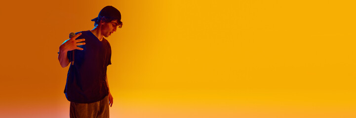 Stylish young man, rap musician in cap and black clothes, holding microphone over yellow background in neon. Concept of music, performance, festival, live concert. Banner. Empty space to insert text