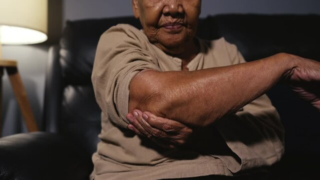 senior woman suffering from elbow pain while sitting on a sofa in living room at night