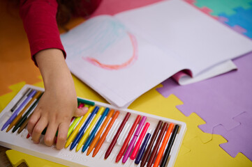 Details on the hands of a little girl taking out colorful pencil from pencil case, drawing beautiful cloud with rainbow, lying on a multi colored puzzle carpet in cozy home interior