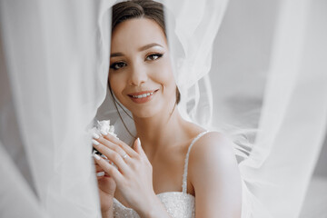 A woman is smiling and holding a flower in her hand. She is wearing a wedding dress and is posing...