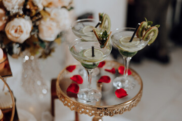 Three martinis with lime slices and kiwi slices on top are on a tray. The tray is on a table with a vase of flowers in the background. Scene is elegant and sophisticated