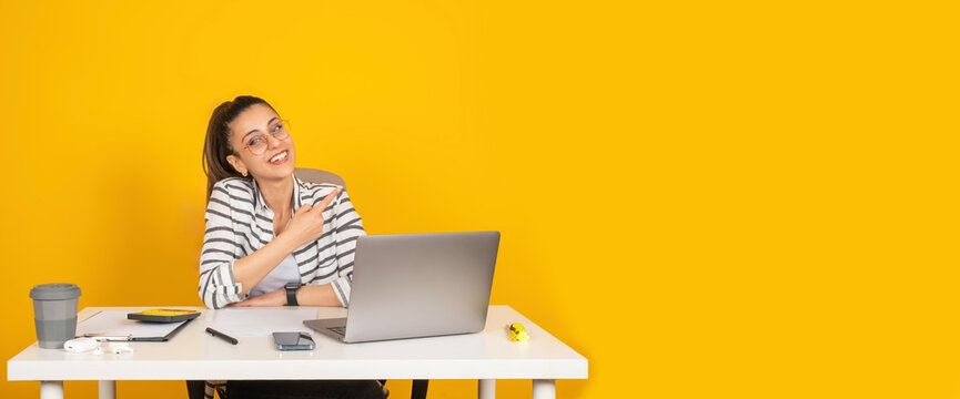 Pointing finger copy space, young happy office worker employee business woman sit office desk pointing finger copy space aside. Yellow background. Career concept. Advertising banner design element.
