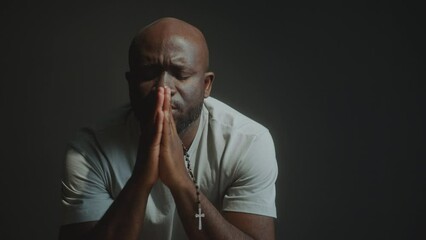 Faithful Black man holding rosary beads with cross and praying deeply to God with palms together and eyes closed
