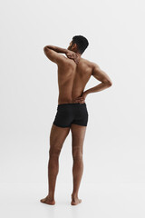 Fototapeta premium Rear view of young African-American shirtless man holding neck and back, standing in underwear isolated on white background. Health care. Concept of male beauty, sport, body care, health, fitness