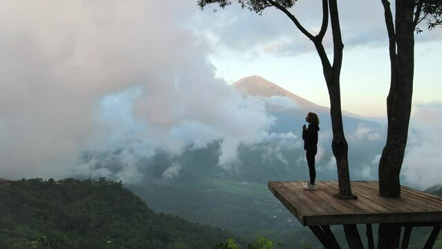 Silhouette of a young girl standing on a wooden platform high in the mountains, folding her hands in namaste and meditating, with a beautiful view of the clouds and the volcano mountain. Bali.