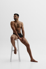 Full-length image of handsome young African-American bearded man sitting on chair shirtless in boxers isolated on white background. Concept of male beauty, sport, body care, health, fitness