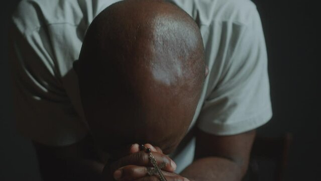 Faithful African American man holding cross and praying deeply with the Bible in times of despair. Tilt-down shot