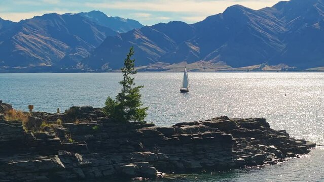 Aerial trucking pan around rocky island reveals sailboat calmly floating in middle of lake