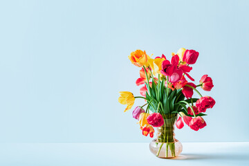 Obraz premium Big flowers bouquet of multicolored tulips in vintage glass vase on light blue background with copy space. Business card. Invitation postcard. Place for greeting text. International holiday. Banner