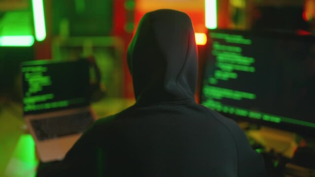 Hacker writing code cracking computer. Man programmer in hoodie cracks system, using computer in dark room back view. Hacker attack, cybercrime, virus software, dark web, cyber security concept.