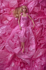 Fototapeta premium The doll lies in a pink plastic sheet and wears a dress made of the same material.Minimal creative fashion concept.Flat lay