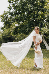 A bride is standing in a field with a white dress and a veil. She is holding a bouquet of flowers and a small handbag. The scene is peaceful and serene, with the bride looking happy and content