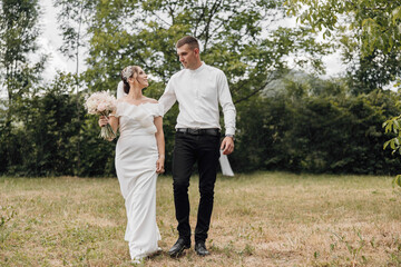 A bride and groom are walking in a field, the bride is holding a bouquet and the groom is holding her hand