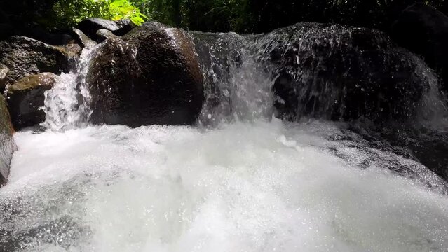 Gentle tracking shot through a picturesque river with a majestic waterfall and large stones, captured in impressive slow motion, 4k resolution