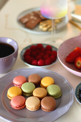 Plate of pastel macarons, cookies and chocolate, cup of tea of coffee, glass of bubble water, various berries, books and accessories on the table. Selective focus, pastel colors.