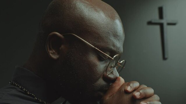Black clergyman in eyeglasses holding hands together and praying to God with eyes closed in church. Close-up shot