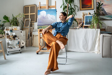 Satisfied dreaming artist woman resting on chair in art studio, smiling looks aside, pondering...