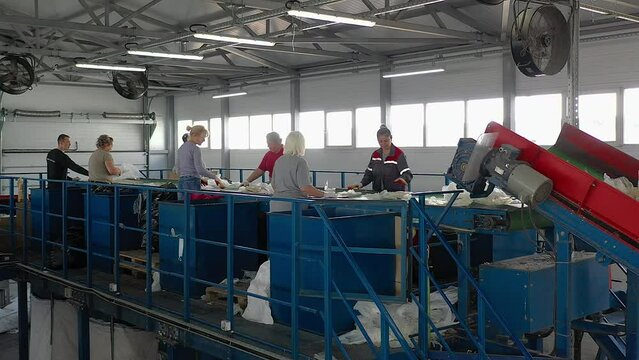 Multiple workers sort plastic at the modern material recycling factory. Workers sort plastic trash coming from the conveyor line. Workers sort various plastic waste into blue boxes. Environment.