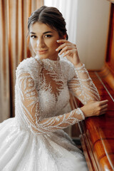 A woman in a wedding dress sits on a wooden table. She is wearing a necklace and has her hand on...