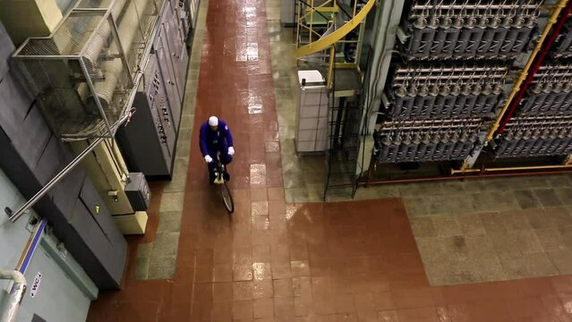 Industrial Manufacturing. A Worker Wearing a Blue uniform and a hat is Riding a Bicycle around the Narrow Hallways of the Workshop Room that is Filled with Gas Centrifuges. Production Process.