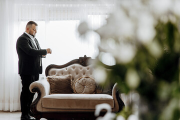 A man in a suit is standing in front of a couch, looking at his watch. The couch is brown and has a...