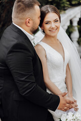 A bride and groom are posing for a picture. The bride is wearing a white dress and the groom is wearing a black suit. They are both smiling and looking at the camera