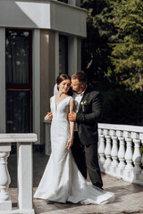 A bride and groom are standing on a balcony, with the bride wearing a white dress and the groom wearing a black suit. They are embracing each other, and the mood of the image is one of love