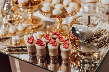 A table with a variety of desserts, including strawberry shortcakes and apple slices. The desserts are arranged on a glass table, and there are also some apples and a bowl of strawberries