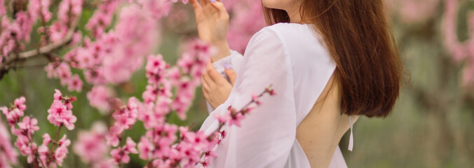 A girl is walking through a field of pink peach flowers. She is wearing a white dress and carrying...