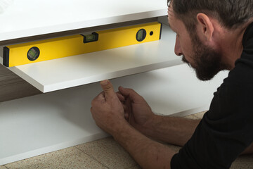 A man installing a wooden shelf on a white wall. A yellow spirit level rests on the shelf ensuring its evenness. They are using both hands to adjust the shelf position. Process of installing shelf