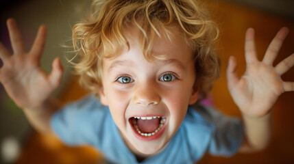 A joyful close-up portrait of a young child with curly blonde hair, wide open blue eyes, and a big smile, reaching towards the camera.
