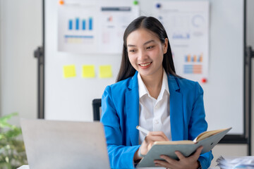 Happy young Asian businesswoman sitting at desk and take notes with laptop computer in the office.
