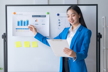 Asian businesswoman pointing at a white board with graphs on it. She is smiling and she is giving a...