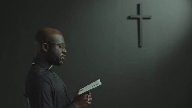 African American priest wearing cassock reading Bible and preaching in modern catholic church with minimalist cross on grey wall. Side view, copy space