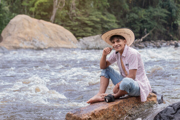 lifestyle. young latin guy wearing a hat sitting on a rock by the river and smiling