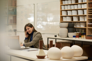 Smiling ceramics studio manager talking on a phone and using a laptop