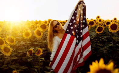 Beautiful young woman with the American flag in a sunflower field at sunset. 4th of July....