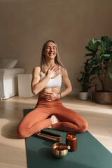 Joyful happy carefree woman smiling, holding hands at heart and belly practicing meditation pranayama on a yoga mat at home on a sunny day, wellness, mental health. 