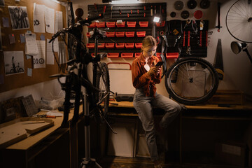 Young woman looking on phone searching help on repairing damaged bike in workshop or garage. Lady...