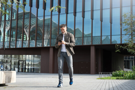 Mature hispanic senior business man in casual clothes using smartphone cellphone app walking at office building. Full body of Latin middle age freelance businessman holding mobile cell phone outdoors