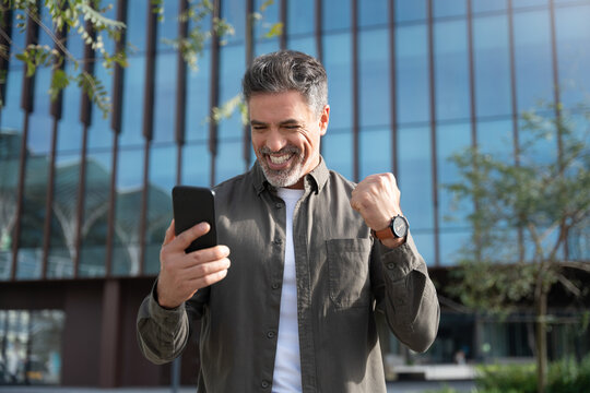 Happy delighted satisfied business man looking smartphone mobile phone gesturing yes winning pose standing at urban city office. Overjoyed excited businessman, doing winner gesture, reading good news
