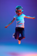 Obraz premium Schoolboy in casual attire jumping of joy while dancing during listening music in headphones in mixed neon light against blue background. Concept of music, dance, fashion and style, technology. Ad