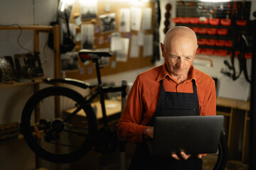 Elderly male mechanic working in a workshop researches information on his laptop